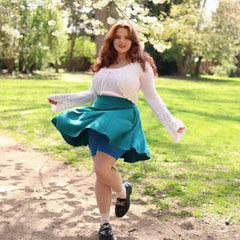 Woman in a white top and teal skirt standing in a park with trees and grass in the background