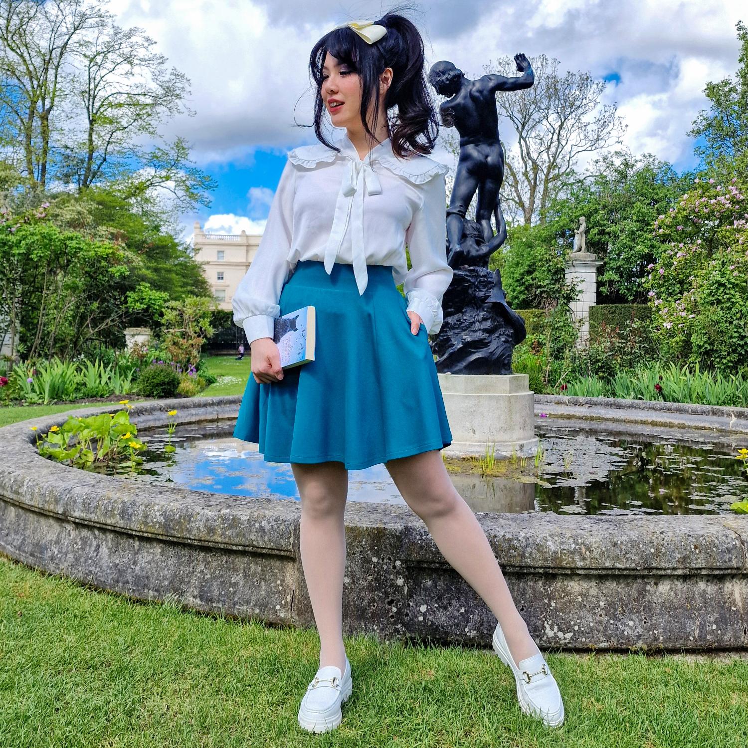 Woman in a white blouse and teal blue skirt standing in a garden with statues and a pond.