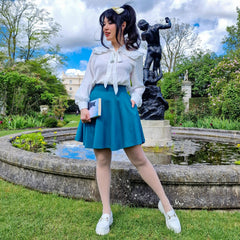 Woman in a white blouse and teal blue skirt standing in a garden with statues and a pond.