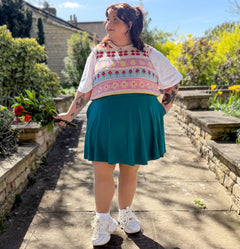 Woman in a garden wearing a colorful sweater and teal skirt, holding flowers.